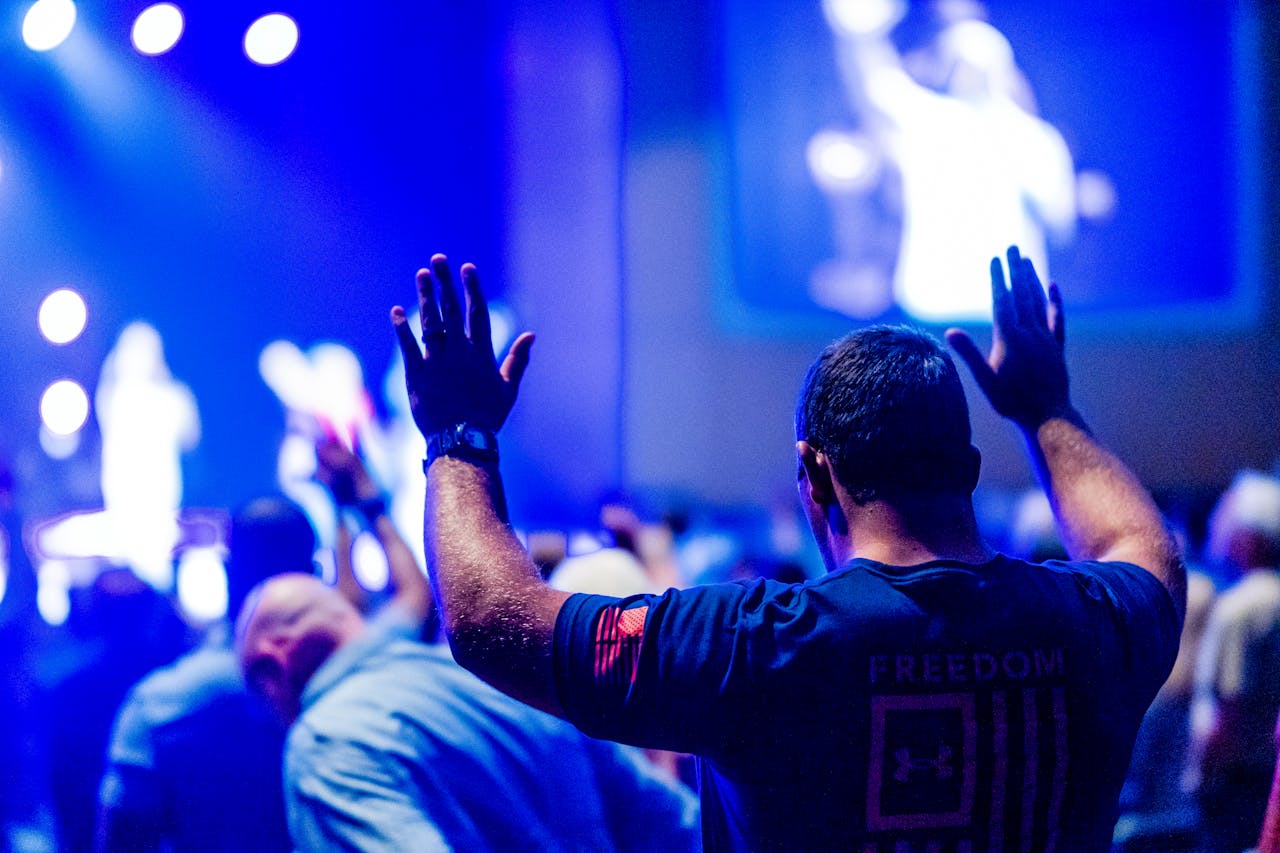 creative-02 A man raises his hands in worship at a vibrant indoor event with a blurred stage.